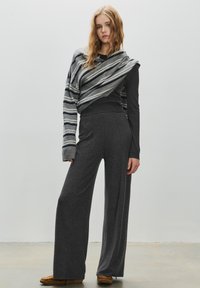 Pantaloni grigi a gamba larga con una texture morbida; abbinati a un top a maniche lunghe scuro. Un maglione a righe oversize drappeggiato sulle spalle.