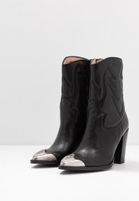 Černé kožené kotníkové boty s špičatou špičkou, zdobené stříbrnými akcenty, embosovaným švy a vysokým blokovým podpatkem. Zapínání na zip.