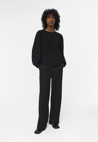 Blusa negra de manga larga con hombros fruncidos, combinada con pantalones de pierna ancha negros de talle alto y mocasines negros brillantes sobre un fondo blanco.