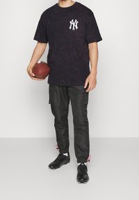 Černé tričko s grafickým potiskem s bílým logem, v kombinaci s černými cargo kalhotami a černými kopačkami. Osoba drží hnědý fotbal.