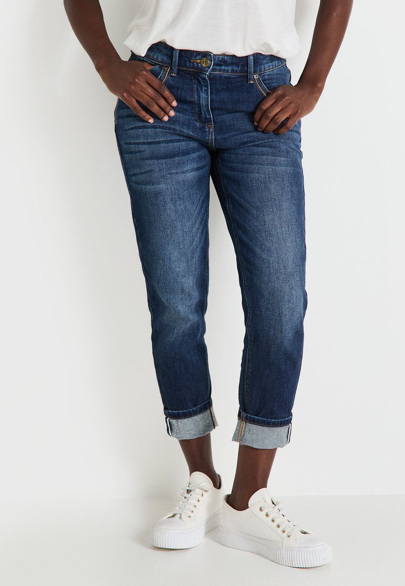 Jean en denim bleu foncé avec une coupe décontractée, ourlets retroussés et poches avant et arrière. Associés à des baskets blanches.