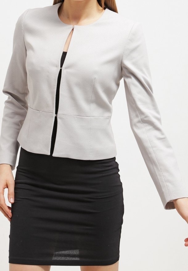 Veste courte gris clair avec un col rond, tissu texturé et fermeture éclair centrale. Portée sur une robe noire ajustée.