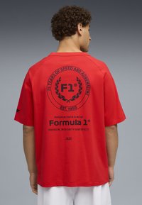 Rød T-shirt med korte ærmer og afslappet pasform. Har sort Formel 1-logo og tekst på ryggen, glat bomuldsstof og rund halsudskæring.