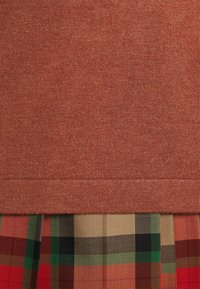 Tissu tricoté couleur rouille au-dessus d’un textile à carreaux multicolores avec des motifs verts, noirs, beiges et rouges.
