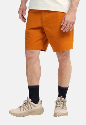 Shorts orange pour homme avec une texture lisse, des poches latérales et une coupe décontractée, associés à des chaussures de sport beige et des chaussettes marine.