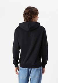 Felpa nera con una texture morbida, dotata di cappuccio con coulisse, polsini e orlo a coste. Indossata con jeans azzurri, vista da dietro.