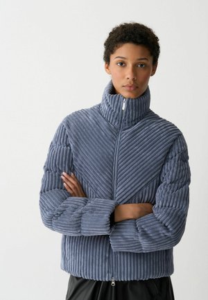 Gewatteerde jas van blauwe corduroy met een hoge kraag, diagonale ribbeltextuur en ritssluiting. Kenmerkt zich door gedurfde horizontale en verticale lijnen.