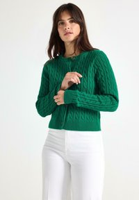 Woman wearing a green cable-knit cardigan and white pants, standing against a plain light background, adjusting the cardigan sleeves.