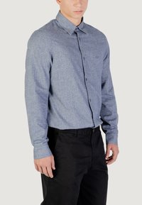 Camicia azzurra chiara con bottoni realizzata in tessuto testurizzato, dotata di un colletto classico e maniche lunghe. Indossata con pantaloni neri.