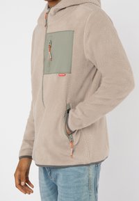 Homme portant une veste polaire beige avec des poches zippées vert clair sur la poitrine et les côtés, assortie à un jean bleu clair, sur fond blanc.