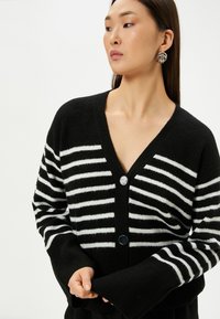 Schwarz-weiß gestreifter Cardigan mit V-Ausschnitt, zwei Knöpfen und weiten Ärmeln; weiche Textur.