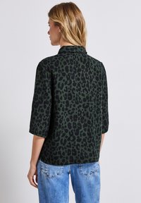 Grøn dyreprint skjorte med korte ærmer, der har en krave og en løs pasform. Texturen virker glat, parret med blå denim jeans.