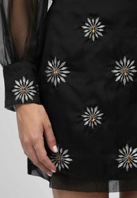 Robe noire avec broderie de marguerites argentées et accents ambres sur les manches transparentes et l'ourlet, mettant en valeur des motifs et textures complexes.