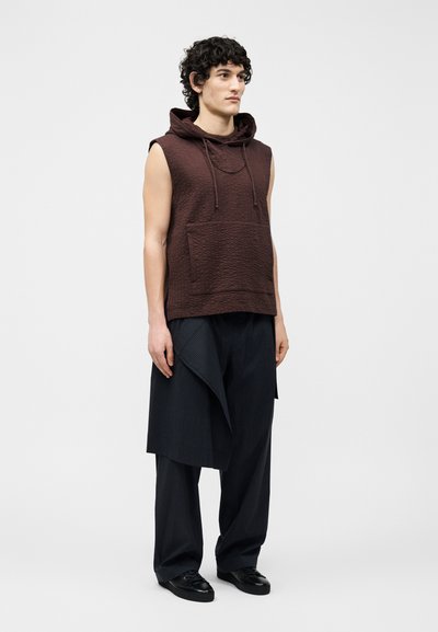 Jonge man met krullend haar, gekleed in een mouwloze bruine hoodie en losse zwarte gelaagde broek, staand tegen een effen lichte achtergrond.