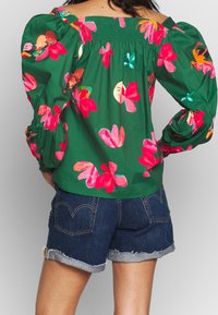 Blouse verte à motifs floraux avec des fleurs roses vives, col smocké et manches bouffantes oversize, associée à un short en jean avec bords effilochés.