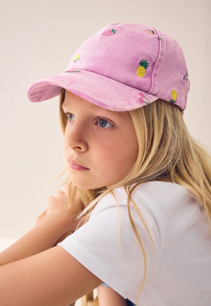 Fille blonde portant une casquette rose avec des fruits brodés, regardant de côté, reposant ses bras sur une surface, portant une chemise blanche à manches courtes.