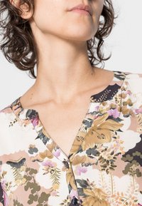 Blusa com padrão floral em tecido suave, com decote em V. Apresenta botões e acabamentos em contraste. As cores incluem bege, verde, rosa e preto.