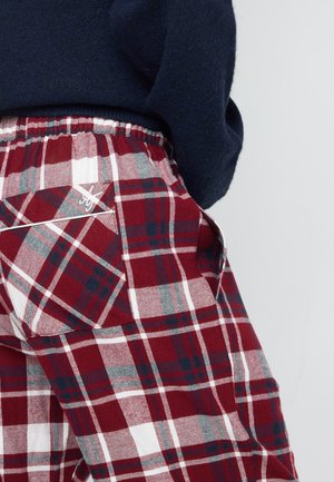 Persoon in rode, witte en donkerblauwe geruite broek met een achterzak en een donkerblauwe trui tegen een effen achtergrond.