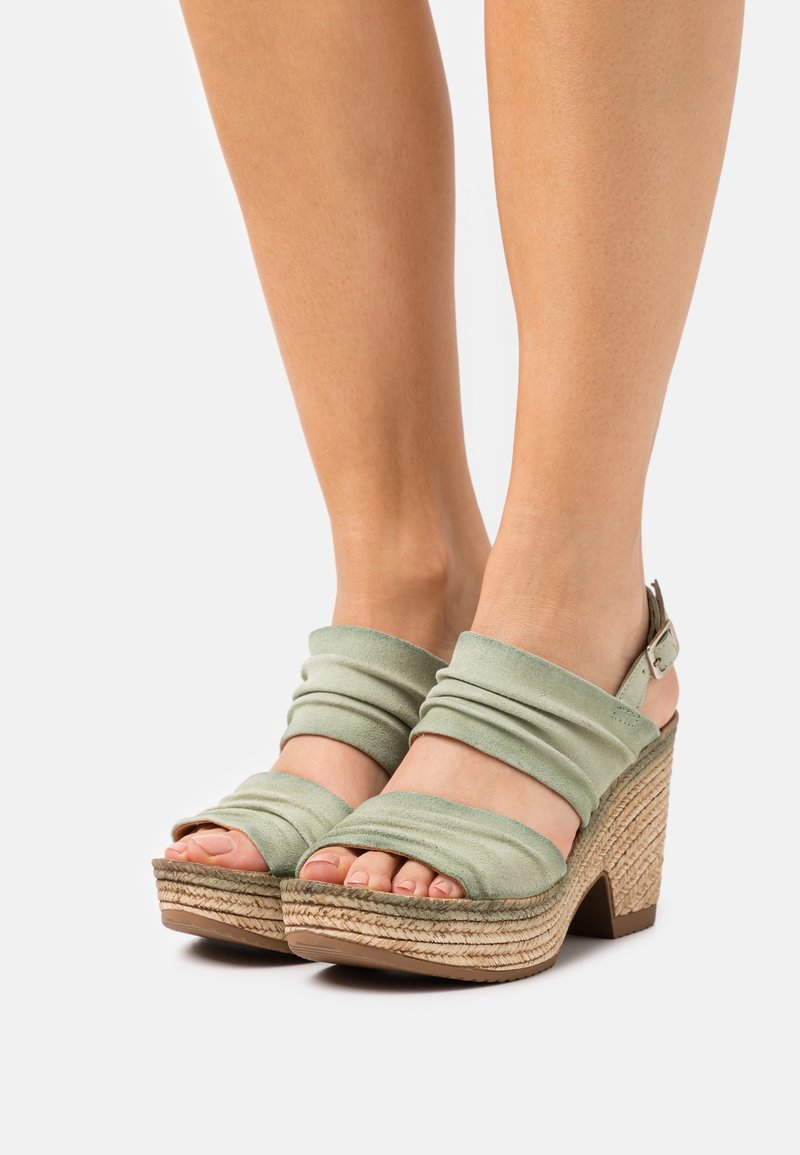 Sandales compensées avec des brides froncées vert clair et des talons plateforme tissés, portées sur des pieds avec un fond neutre.