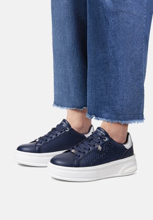 Meripati kulit navy dengan detail berlubang, sol platform putih, penutupan tali, dan aksen perak, dipadukan dengan denim robek.