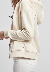 Krémová mikina s kapucí a zipem vpředu, se žebrovanými manžetami a malým zeleným logem listu na spodním lemu. Měkce tkaná látka s hladkým povrchem.