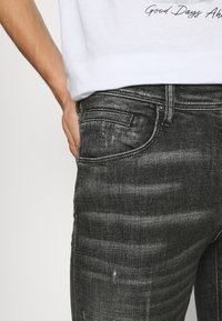 Jeans de denim negro con una leve textura desgastada, diseño de cinco bolsillos, un sutil desvanecimiento y detalles en metal plateado.