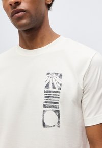 Άνδρας που φοράει εκρού μπλουζάκι με μαύρο αφηρημένο κάθετο σχέδιο και κείμενο "Marc O'Polo EST. IN STOCKHOLM" στο αριστερό στήθος.