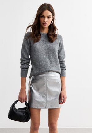 Suéter gris con cuello redondo, combinado con una falda mini metálica plateada. Sosteniendo un pequeño bolso negro, la modelo se queda de pie con el cabello lacio.
