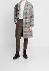 Persoon met een paarse, op maat gemaakte broek, zwarte veterschoenen, een witte coltrui en een grijs geruit, dubbelzijdig lang jasje met zakken, hand in de zak.