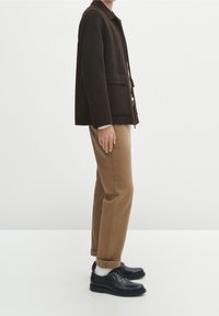 Osoba stojąca w czarnej kurtce, beżowych spodniach z podwiniętymi nogawkami, białych skarpetkach i czarnych skórzanych butach zapinanych na sznurówki na białym tle.