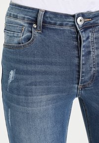 Närbild av blå denimjeans med en knapp och dragkedja, med subtila slitningar nära framfickan och sömmarna.
