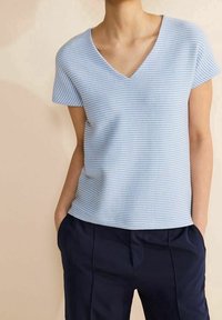 Ljusblus topp med kort ärm och V-ringning. Tyget har horisontella ränder och en mjuk, texturerad yta. Samman med marinblå byxor.