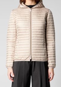 Veste matelassée beige avec motif matelassé horizontal, fermeture à glissière avant et capuche. Texture lisse et poignets élastiques. Portée sur un vêtement noir.