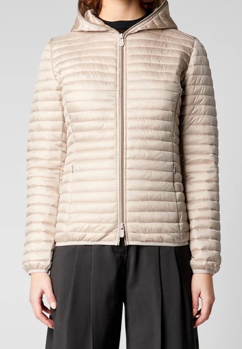 Veste matelassée beige avec motif matelassé horizontal, fermeture à glissière avant et capuche. Texture lisse et poignets élastiques. Portée sur un vêtement noir.