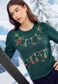 Zeleni pleteni pulover shelinastim izrezom, z božičnimi vezeninami besedila in dreves, poudarjenimi s sijočimi bleščicami, v snežni kulisi.