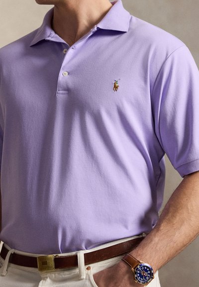 Άνδρας που φοράει μπλουζάκι πόλο λεβάντας με μικρό κεντημένο λογότυπο αλόγου, καφέ δερμάτινη ζώνη, λευκό παντελόνι και μπλε ρολόι χειρός.