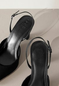 Czarne buty na obcasie typu slingback z teksturowaną cholewką, regulowanym paskiem wokół kostki oraz okrągłym noskiem. Subtelna marka na wkładce i gładkie wykończenie.