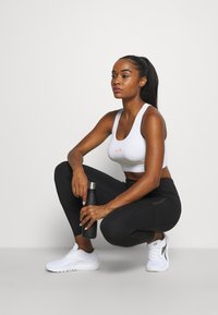 Fehér sportmelltartó narancssárga logóval, fekete leggings oldalzippel, és fehér sportcipő. Ülő pózban fekete vízpalackot tart a kezében.