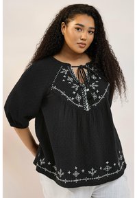 Svart texturerad blus med puffärmar, prydd med vit blommig broderi och en knytkrage. Löst skuren med en dekorativ fåll.