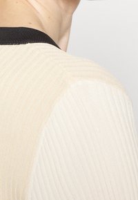 Gros plan sur l'épaule d'une personne portant un pull beige et blanc côtelé avec un col noir, sur un fond gris clair.