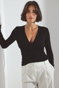 Mujer con cabello corto castaño que lleva una blusa negra de manga larga con escote en V fruncida y pantalones blancos de tiro alto, de pie con un brazo descansando en la pared.