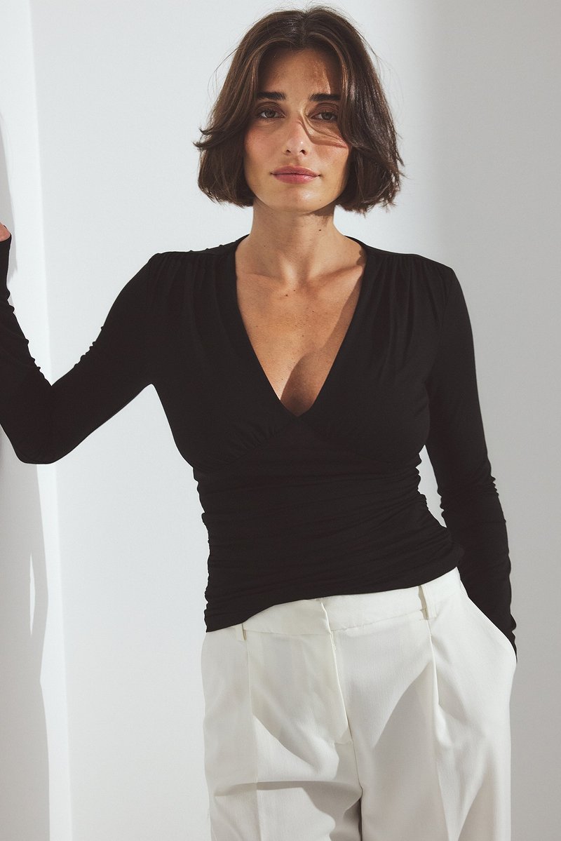 Mujer con cabello corto castaño que lleva una blusa negra de manga larga con escote en V fruncida y pantalones blancos de tiro alto, de pie con un brazo descansando en la pared.