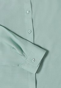 Chemise verte clair à manches longues avec poignets boutonnés et boutons sur le devant, montrant une texture de tissu détaillée et des coutures apparentes.