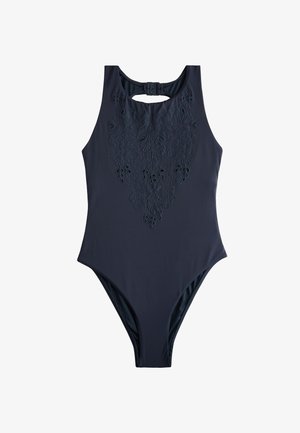 Femme portant un maillot de bain une pièce foncé, debout sur une plage avec les cheveux mouillés, des vagues de l'océan et des rochers en arrière-plan.