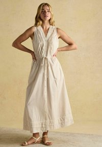 Prúžkované maxi šaty bez rukávov v krémovej a hnedej farbe. Majú gombíkový predný diel, nastaviteľný opasok a nariasený lem. Nosené s plochými sandálmi.