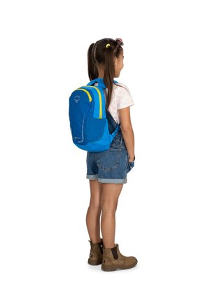 Sac à dos bleu avec des accents jaunes, doté de bretelles rembourrées, d'un tissu texturé et de poches latérales, porté par un enfant en shorts en denim et des bottes marron.
