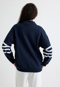 Sudadera azul marino con cuello acanalado. Presenta rayas blancas en contraste en las mangas y un ajuste relajado con hombros caídos.
