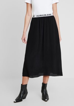 Kobieta nosząca czarną spódnicę midi z białym elastycznym pasem z napisem „Calvin Klein Jeans” w połączeniu z czarnymi skórzanymi botkami za kostkę.