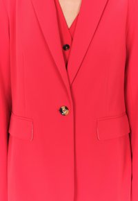 Blazer rosso brillante con tessuto liscio, chiusura con un bottone nero singolo e due tasche frontali. Sottogiacca a V dello stesso colore.