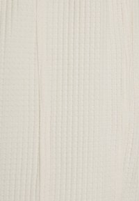 Žebrovaná pletenina v měkké bílé barvě. Vyznačuje se vertikálními liniemi s jemnou strukturou a hladkým povrchem. Bez viditelných vzorů nebo kovových prvků.